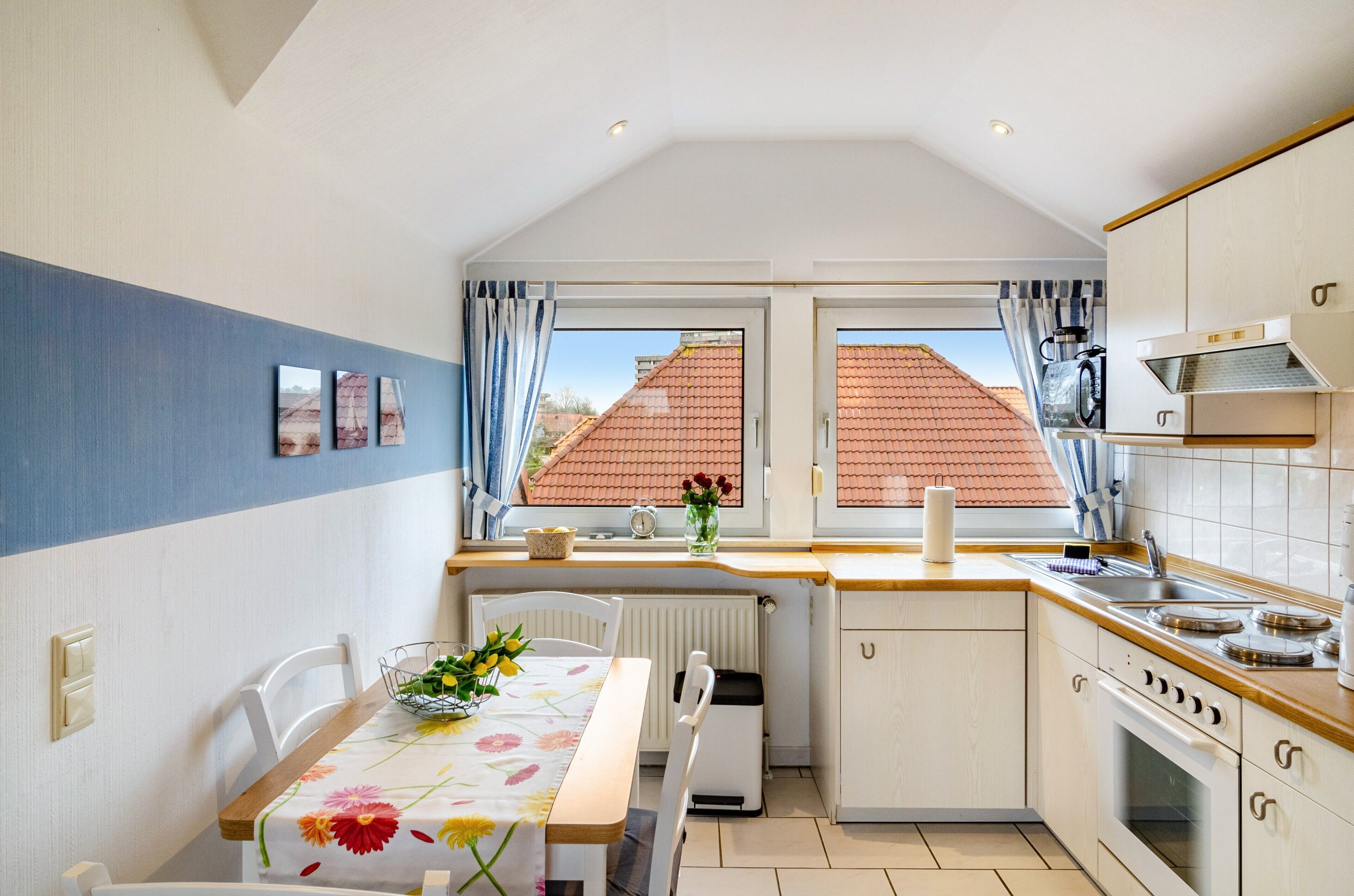 A compact kitchen with a dining area, a radiator, a window with curtains, and a view of a red-roofed building.