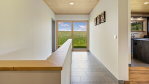Een hal met een houten bankje, uitzicht op een groen veld door een glazen deur, en een keuken met een aanrecht en spoelbak.