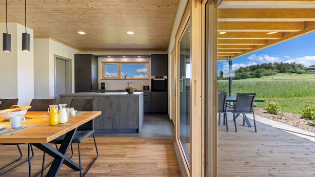 Een moderne keuken met een centraal eiland, een eethoek met een houten tafel en stoelen, en uitzicht op een groen veld door grote ramen.