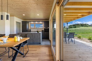 A modern kitchen with a central island, a dining area with a wooden table and chairs, and a view of a green field through large windows. - General description of the vacation apartment at the Hummelberghof (Rosenfeld)
