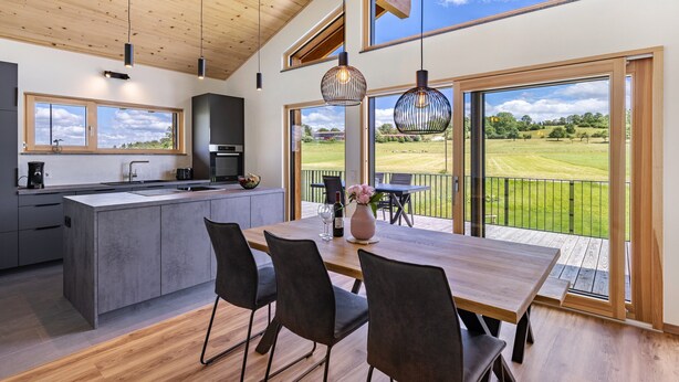 Een moderne keuken met een groot eiland, een eethoek met stoelen en grote ramen met uitzicht op een groen landschap.