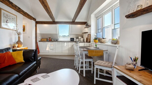 A modern kitchen with white cabinets, a dining area with a white table and chairs, a black leather sofa with colorful cushions, and a large window providing natural light. - Apartment 'Gemütliche Ferienwohnung In Seenähe' with Shared Terrace, Shared Garden and Wi-Fi (Neustrelitz)
