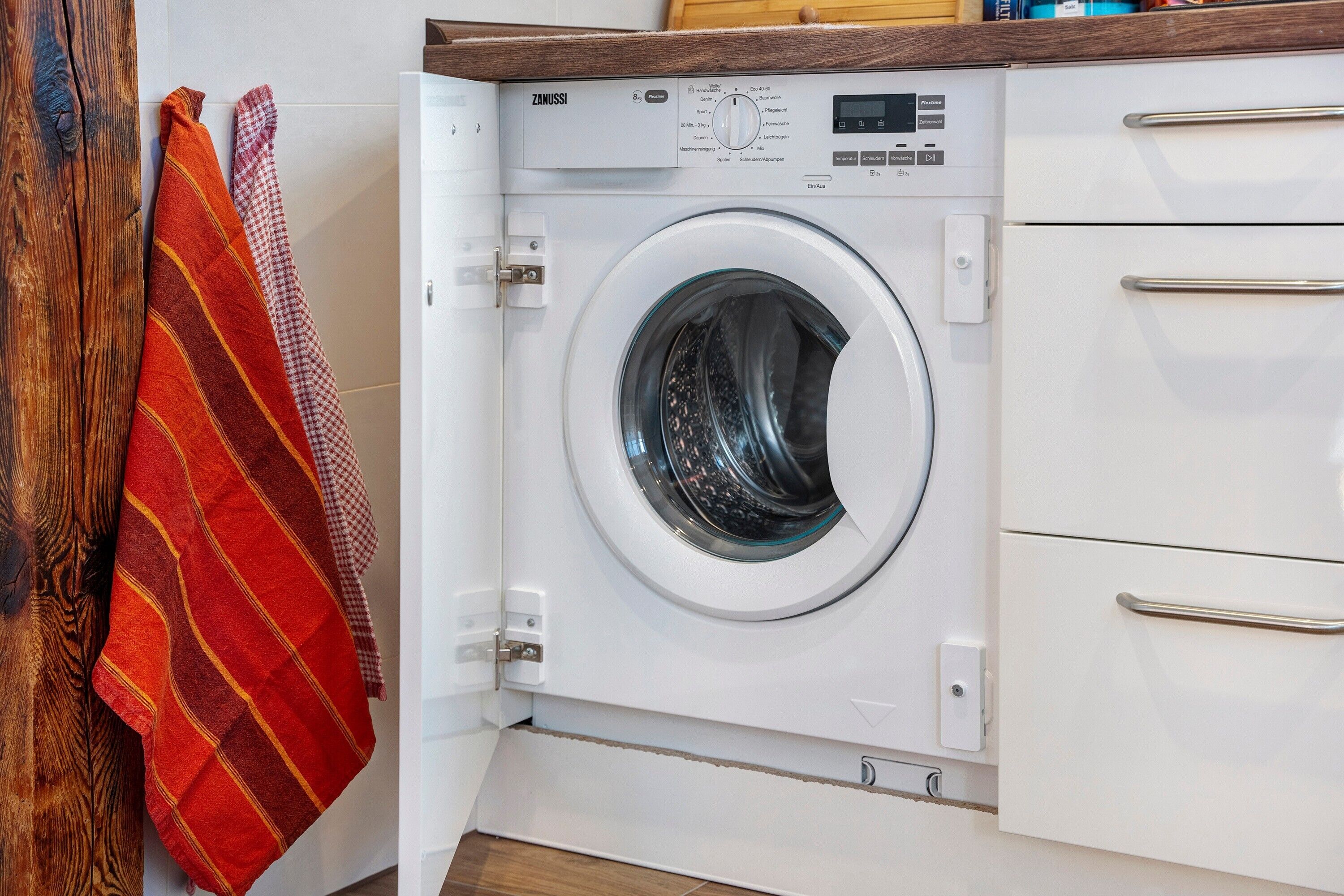 A front-loading washing machine in a kitchen with wooden cabinets and a wooden floor.