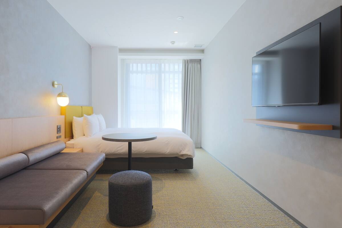 A modern hotel room with a bed, a sofa, a small round table, a television mounted on the wall, and a window with curtains.