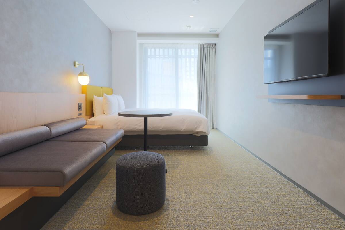 A modern hotel room with a sofa, a round table, a flat-screen TV mounted on the wall, and a large window with curtains.
