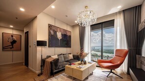 A modern living room with a sofa, a coffee table, and a red chair. There is a painting on the wall and a large window with a view. - The LUMIÈRE Riverside By An Gia Hospitality (Ho Chi Minh City)