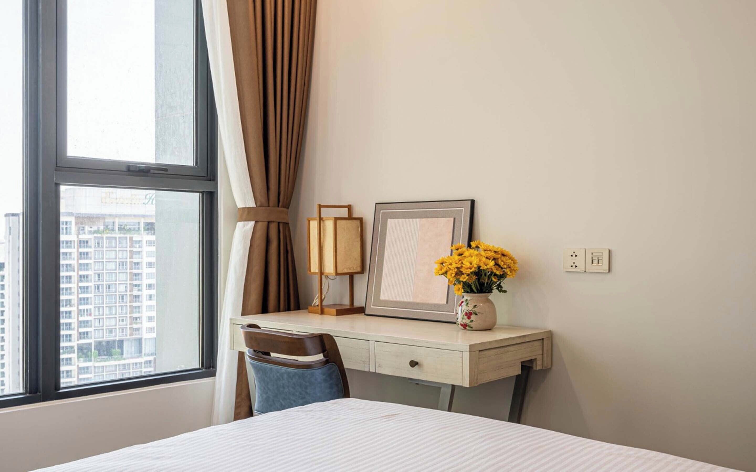 A hotel room with a bed, a desk with a lamp, a vase with flowers, and a framed picture on the wall.