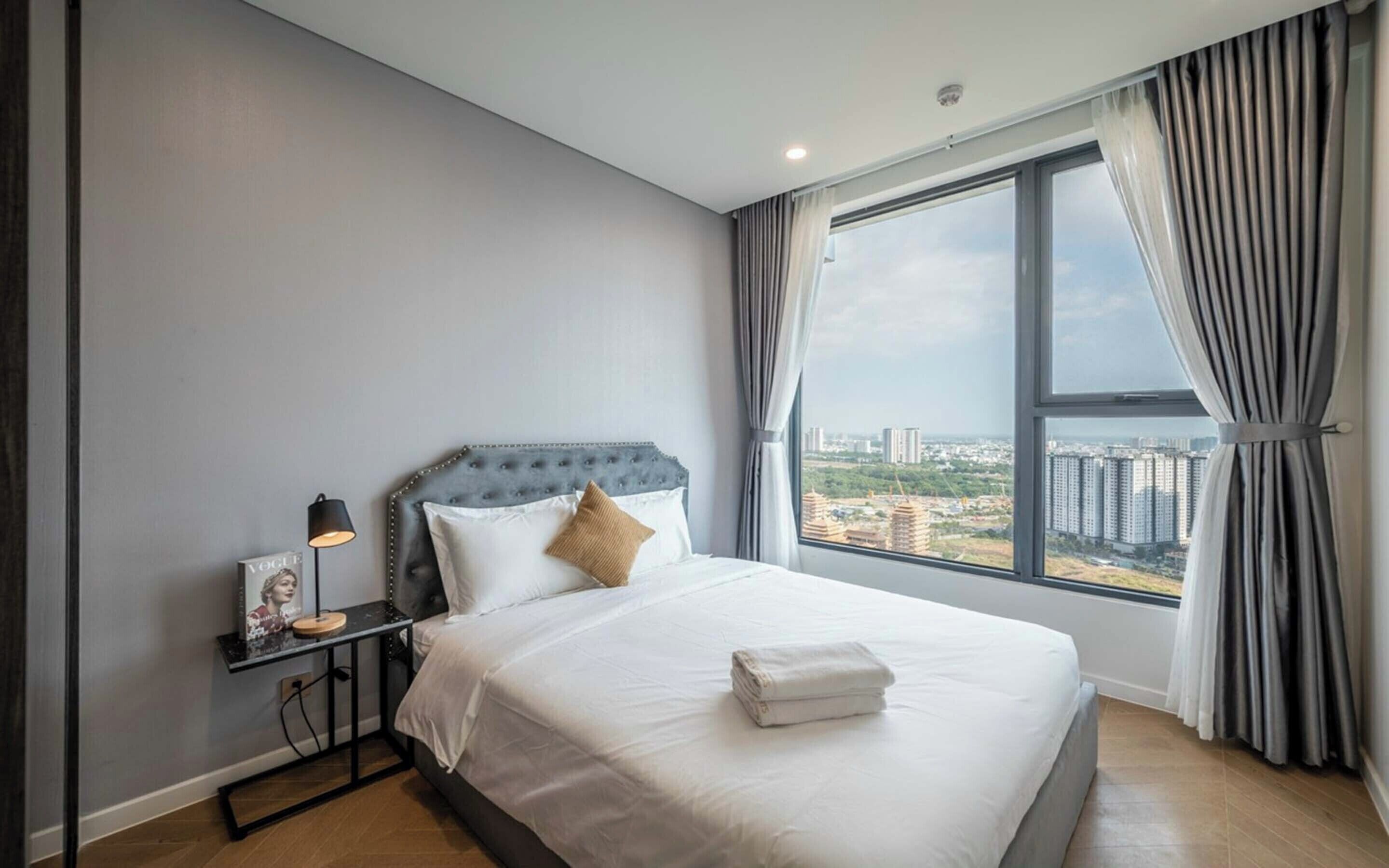 Et moderne hotelværelse med en stor seng, et natbord, en lampe og udsigt over byen gennem et stort vindue.