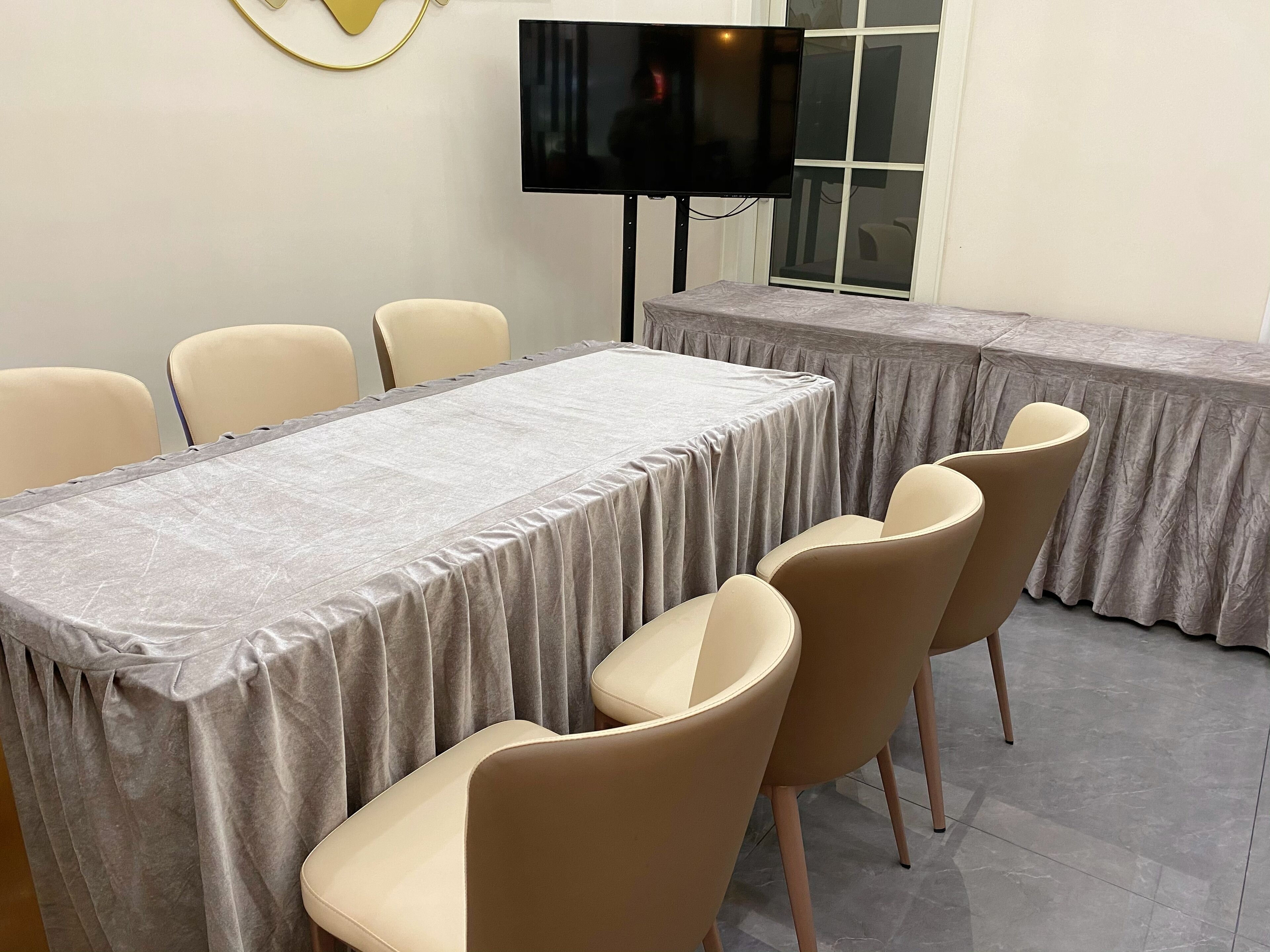 A conference room with a long table covered in a grey tablecloth, surrounded by beige chairs. A flat-screen TV is mounted on the wall above the table.