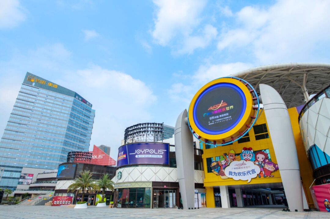 A modern urban plaza with a prominent circular building featuring a digital display and colorful signage.