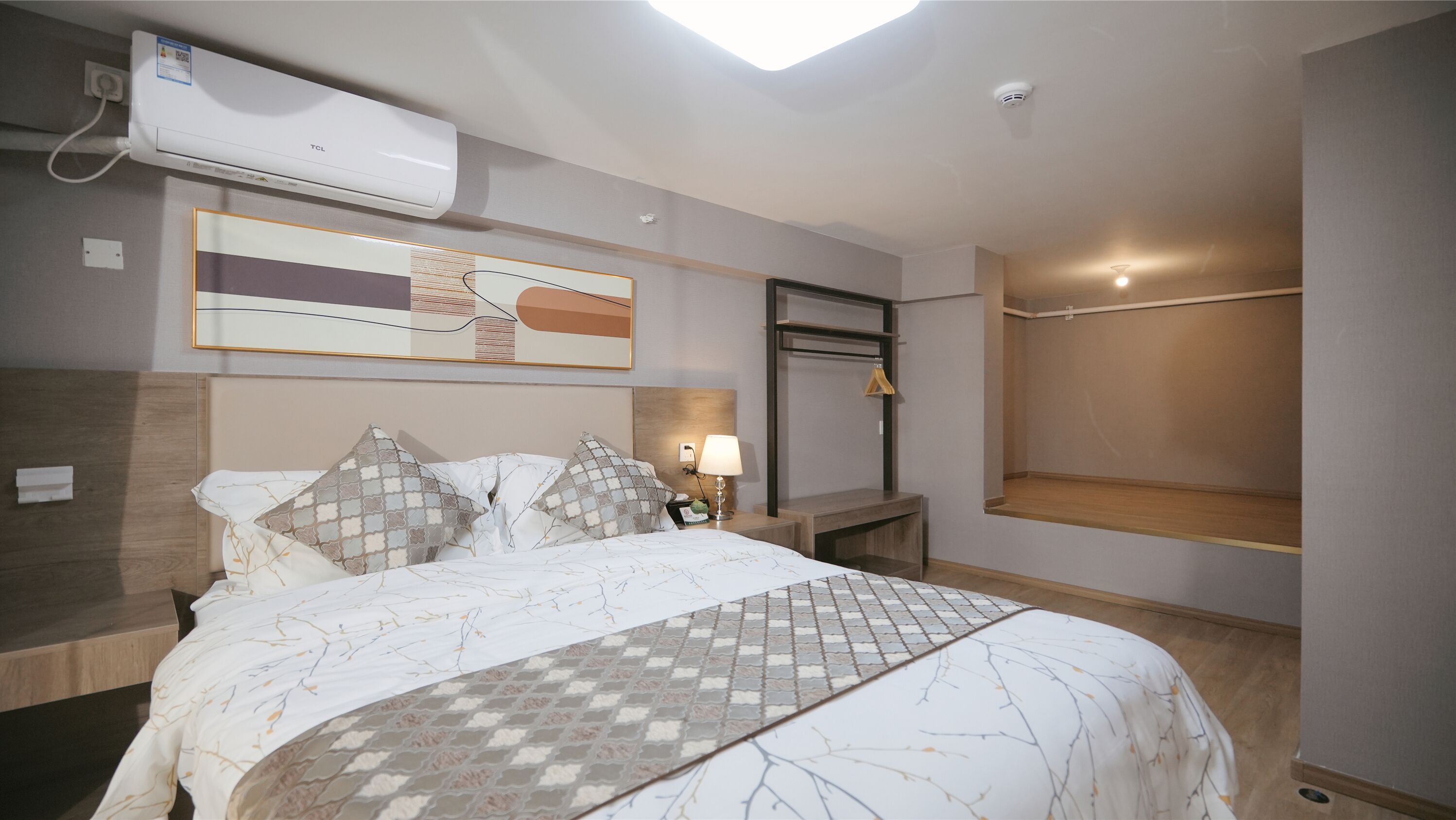 A hotel room with a bed, a nightstand, a wall-mounted air conditioner, a painting, and a wardrobe.