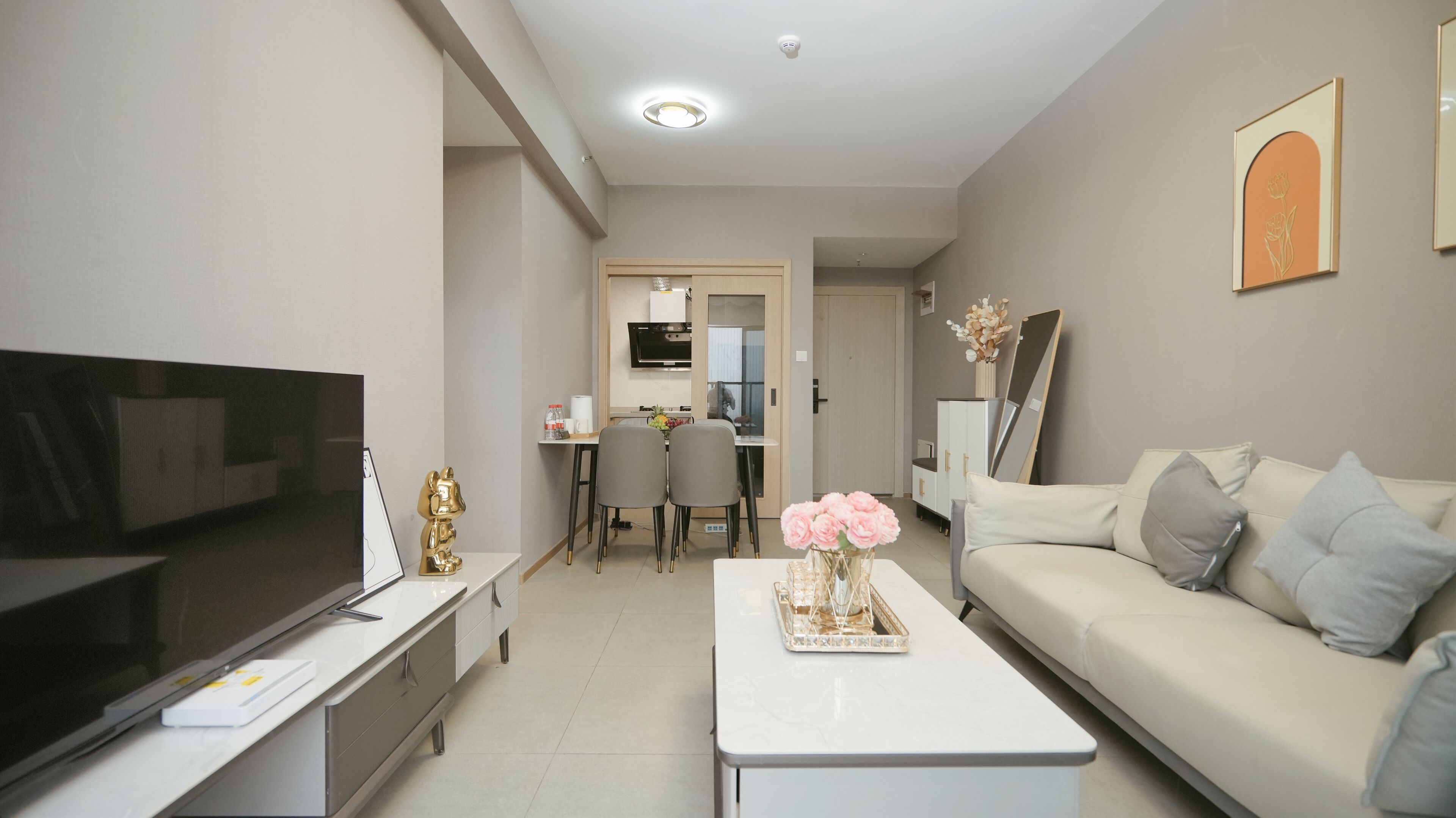 Una sala moderna con un televisor de pantalla plana, una mesa de centro blanca con flores rosadas, un sofá con cojines grises y un comedor con sillas y una mesa.