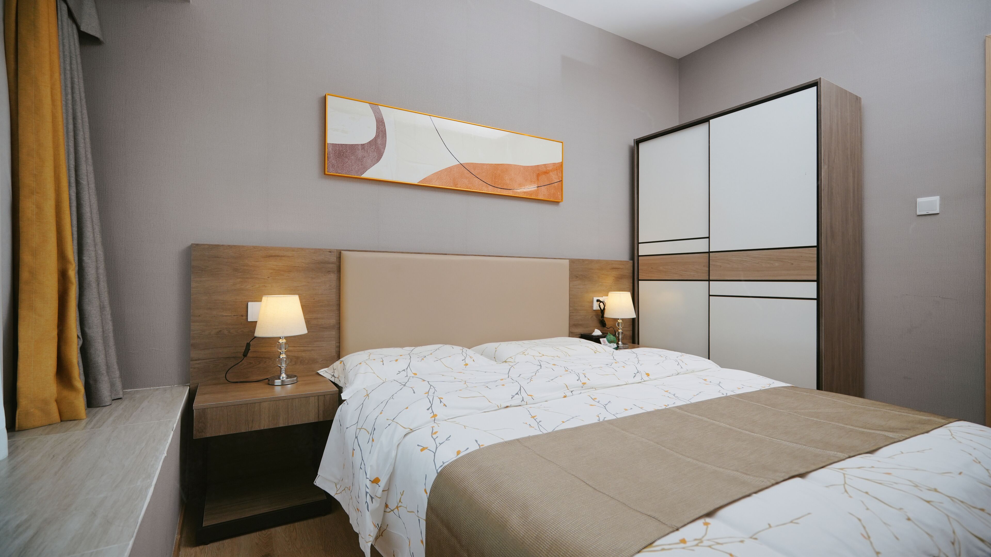 A hotel room with a bed, two bedside tables with lamps, a wooden headboard, a large abstract painting, and a wardrobe.