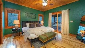 A bedroom with a large bed, wooden flooring, a ceiling fan, and a view of the outdoors through a sliding door.