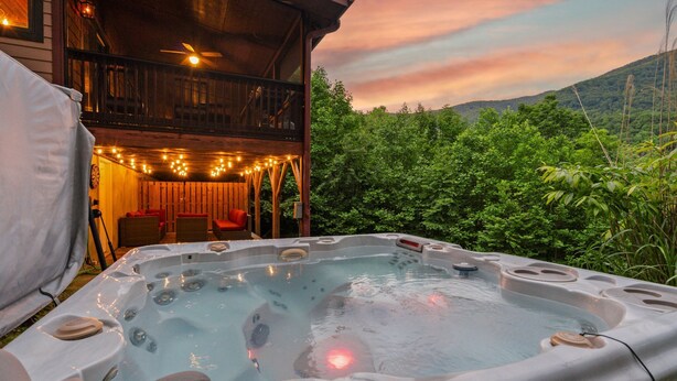 A hot tub with multiple jets, surrounded by lush greenery and a wooden deck with string lights.