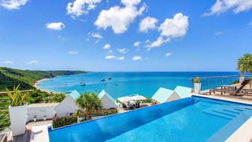 A pool with a view of the sea and boats, surrounded by greenery and a white building with 'Villa Five' signage.