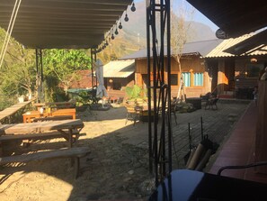 A rustic outdoor seating area with wooden benches and tables, surrounded by potted plants and a mountainous backdrop.