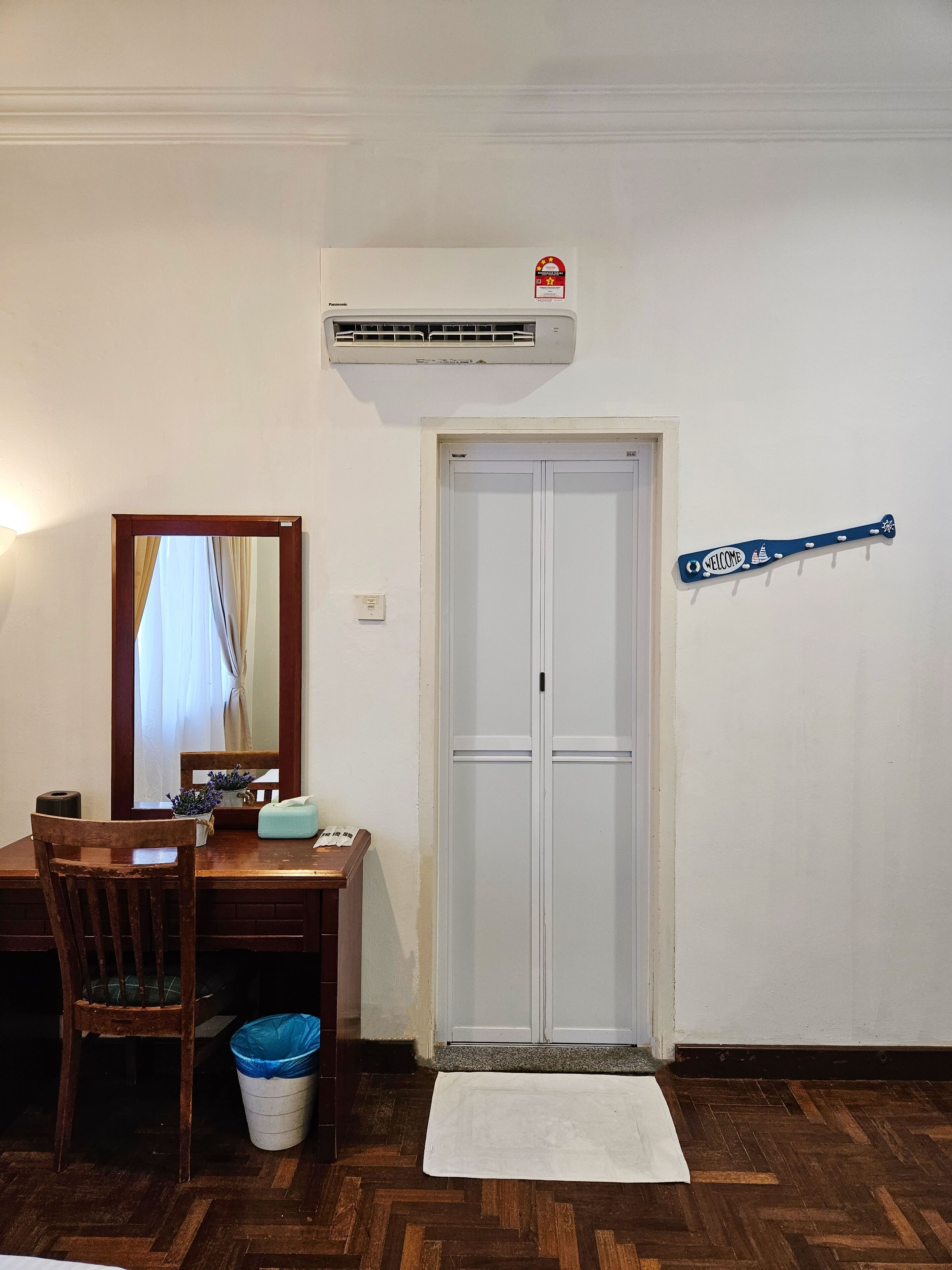 A room with a wooden desk, chair, and a blue trash bin. There is a white door with a 'Welcome' sign, an air conditioning unit above, and a small rug on the floor.