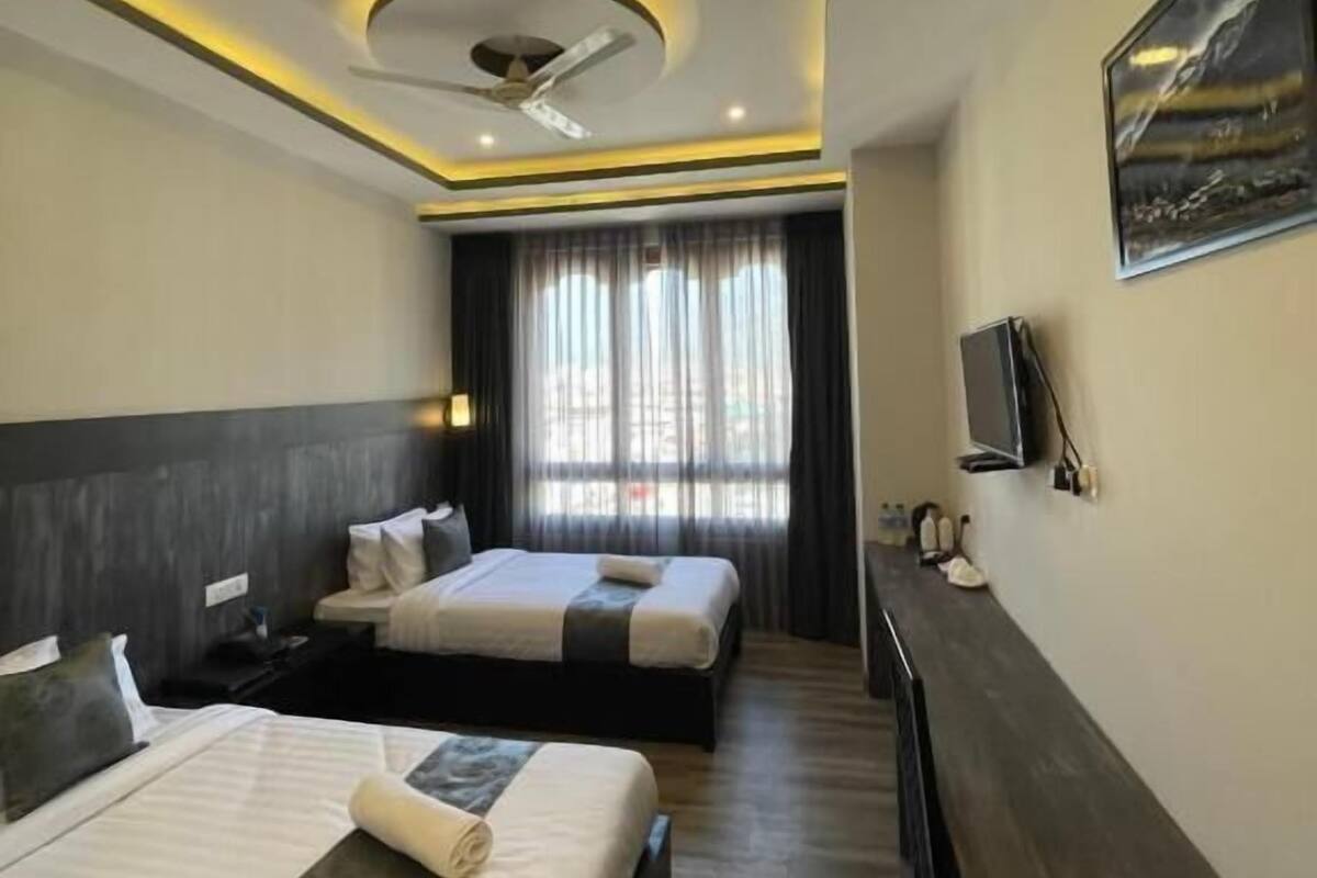 A hotel room with two beds, a desk, a ceiling fan, and a painting on the wall.
