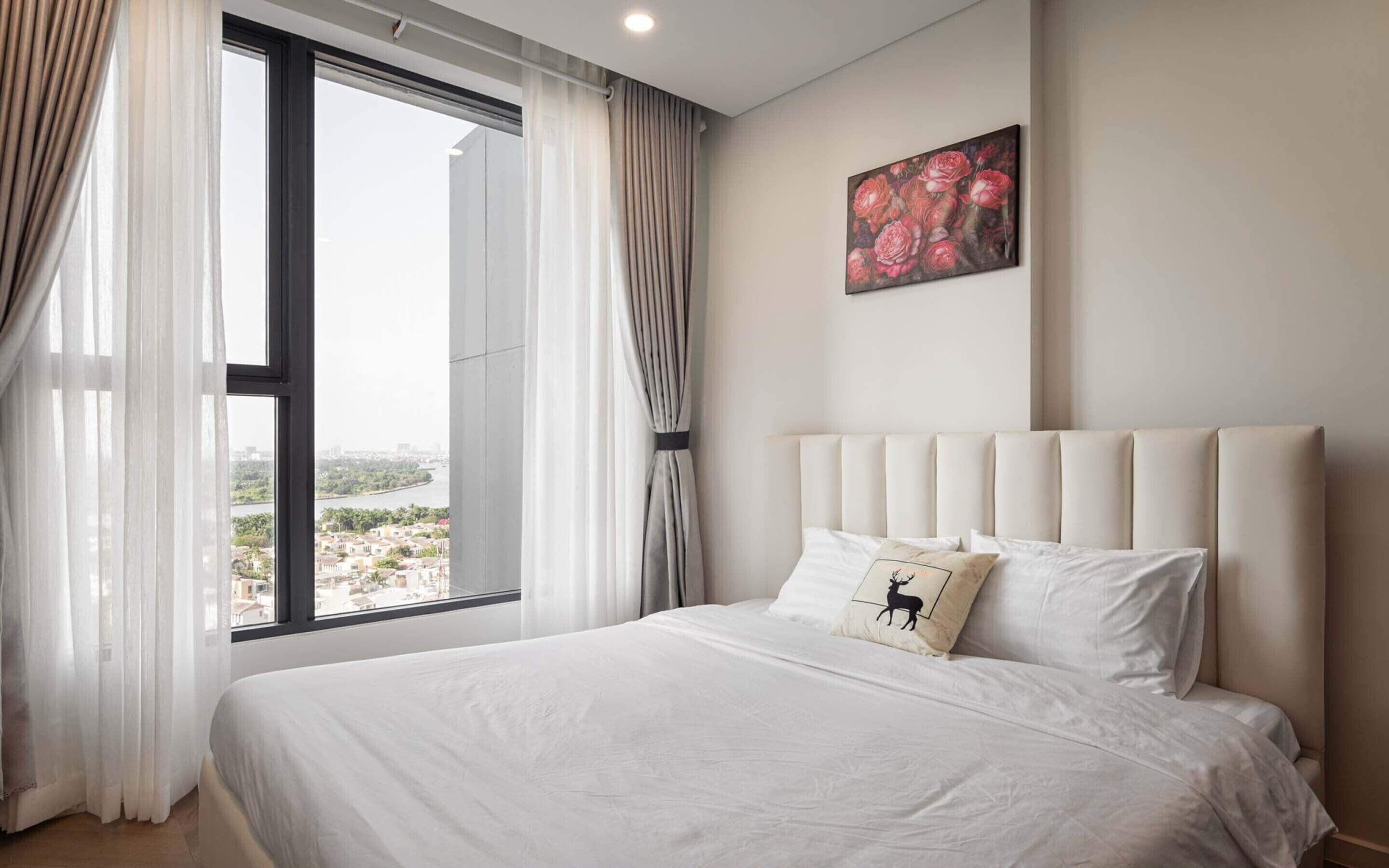 Quarto de hotel com uma janela grande, uma cama com roupa de cama branca, uma cabeceira e um quadro floral na parede.