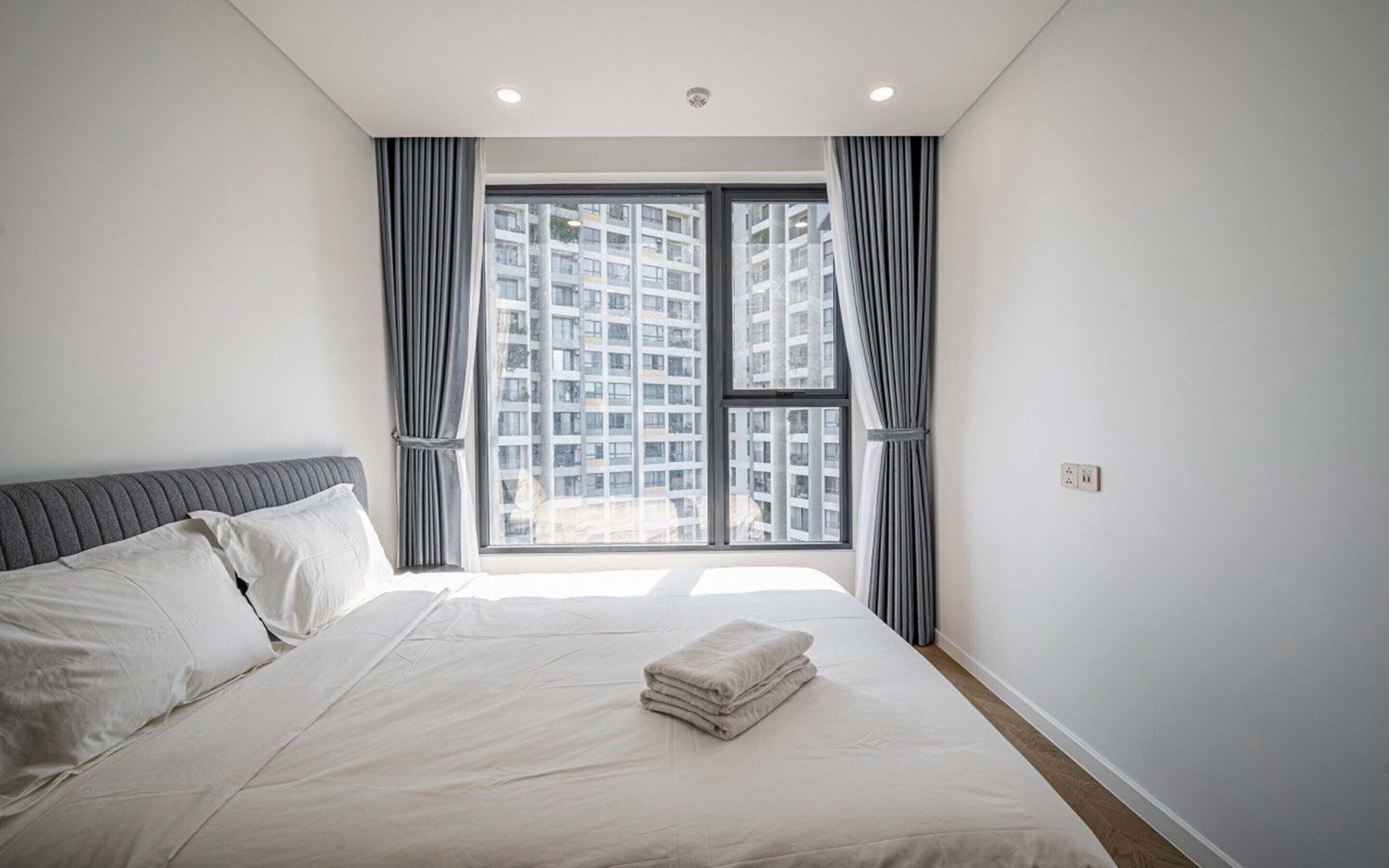 A modern bedroom with a large bed, grey curtains, and a view of high-rise buildings.