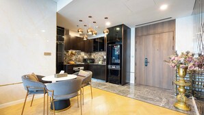 A modern kitchen with dark wood cabinets, a marble countertop, and a built-in oven. There is a dining area with a round table and chairs, and a decorative vase with flowers on the right. - The LUMIERE Riverside Residence (Ho Chi Minh City)