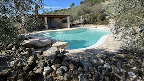 A swimming pool surrounded by rocks and olive trees, with a covered outdoor seating area in the background.