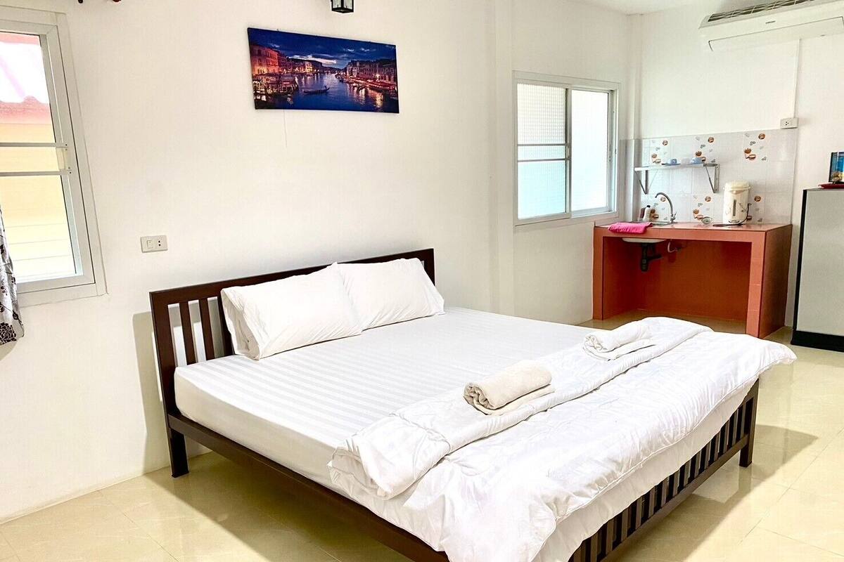 A neatly made bed in a hotel room with a cityscape painting above it, a small desk with a sink, and a window with blinds.