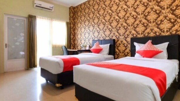 A hotel room with two beds, each with white bedding and red accents, a desk with a chair, and a patterned wallpaper. - Omah Manahan (Surakarta)