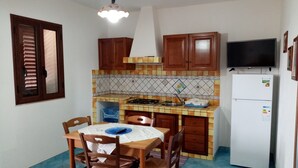 A compact kitchen with wooden cabinets, a gas stove, a sink, and a refrigerator. There is a dining table with chairs and a television mounted on the wall.