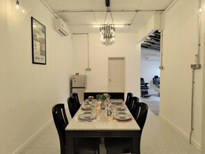 Un coin repas avec une longue table dressée pour six personnes, des chaises noires, un lustre et un collage encadré accroché au mur.