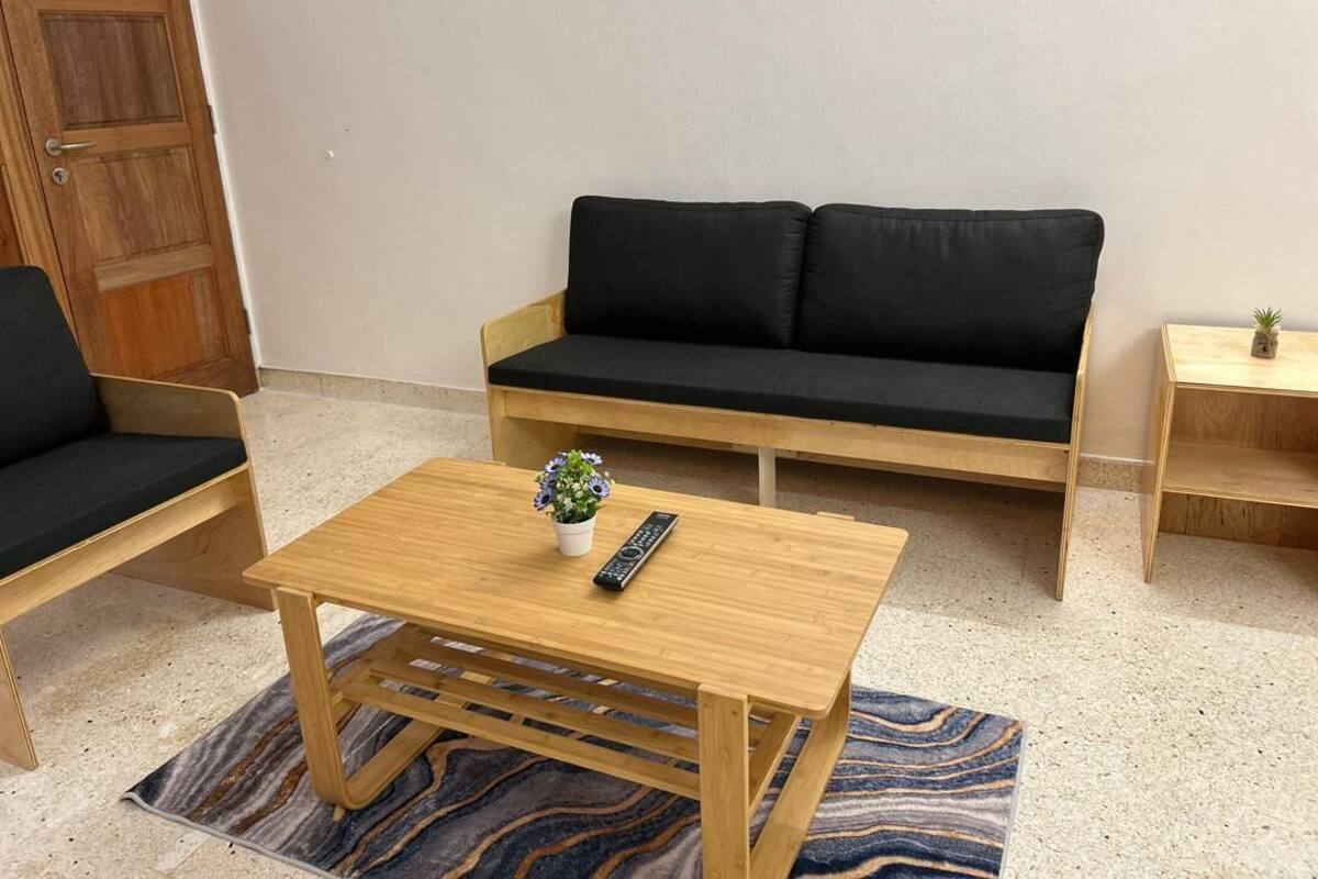 A room with a wooden sofa, a matching wooden armchair, a small wooden table with a remote control and a potted plant, and a patterned rug on the floor.