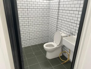 Une petite salle de bain propre, avec des murs et un sol carrelés de blanc, des toilettes blanches et un lavabo blanc avec un flexible.