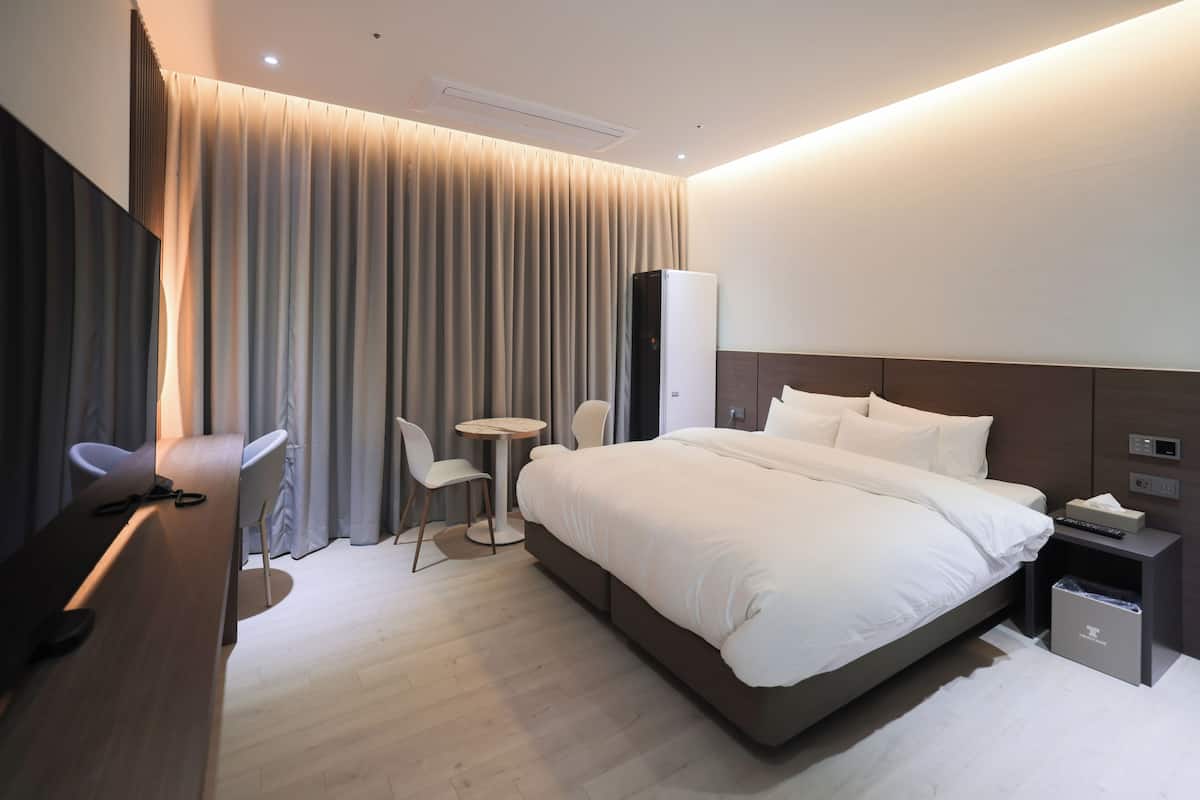 A modern hotel room with a large bed, a desk, a chair, a small round table, and a wardrobe.