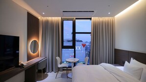 Habitación de hotel con cama, escritorio con computadora, televisión, pequeña mesa con sillas y ventana con cortinas.