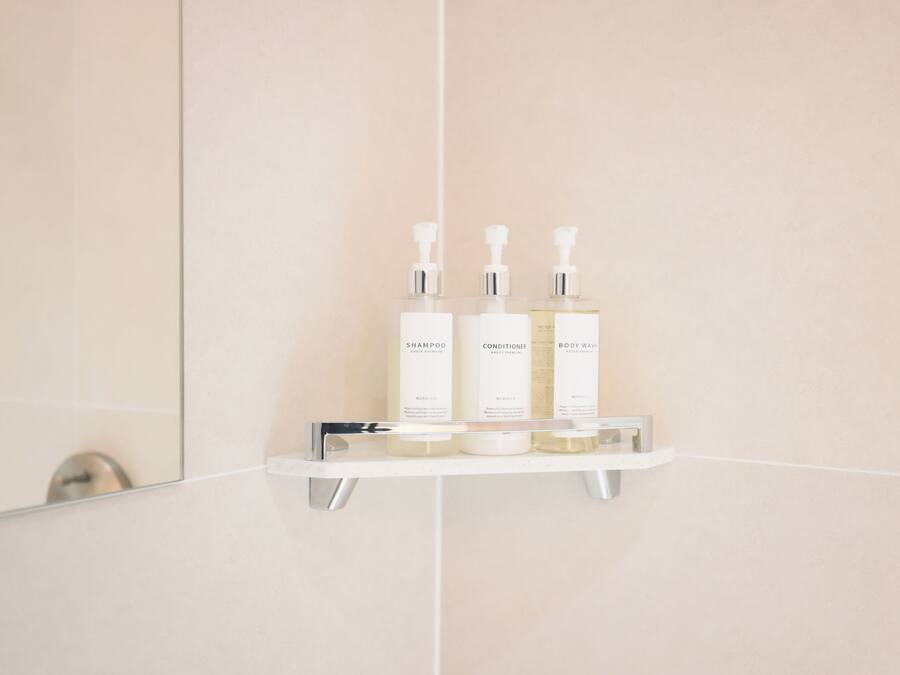 Un baño con un estante blanco que contiene tres botellas de champú, acondicionador y gel de ducha.