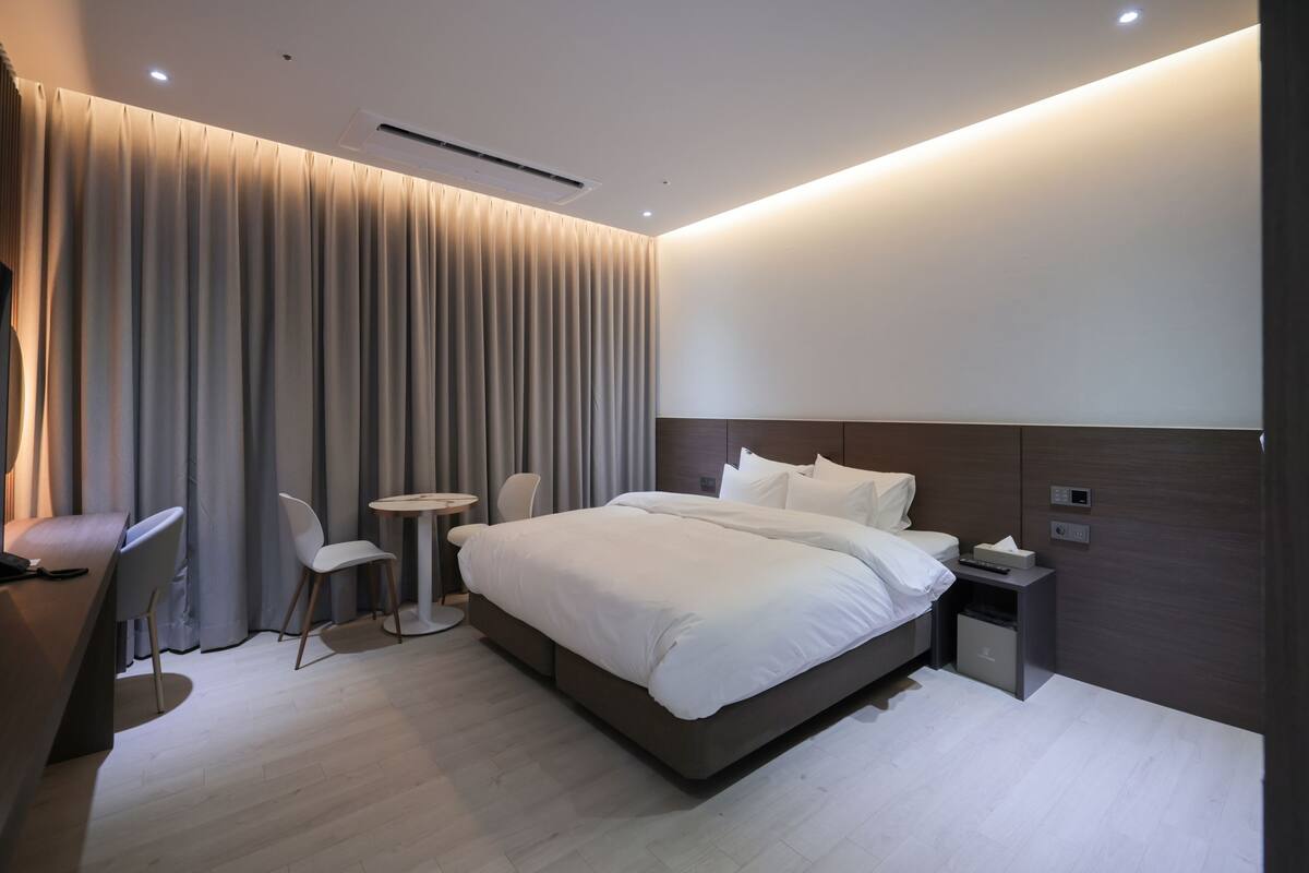 A modern hotel room with a large bed, bedside table, and a small desk with a chair.