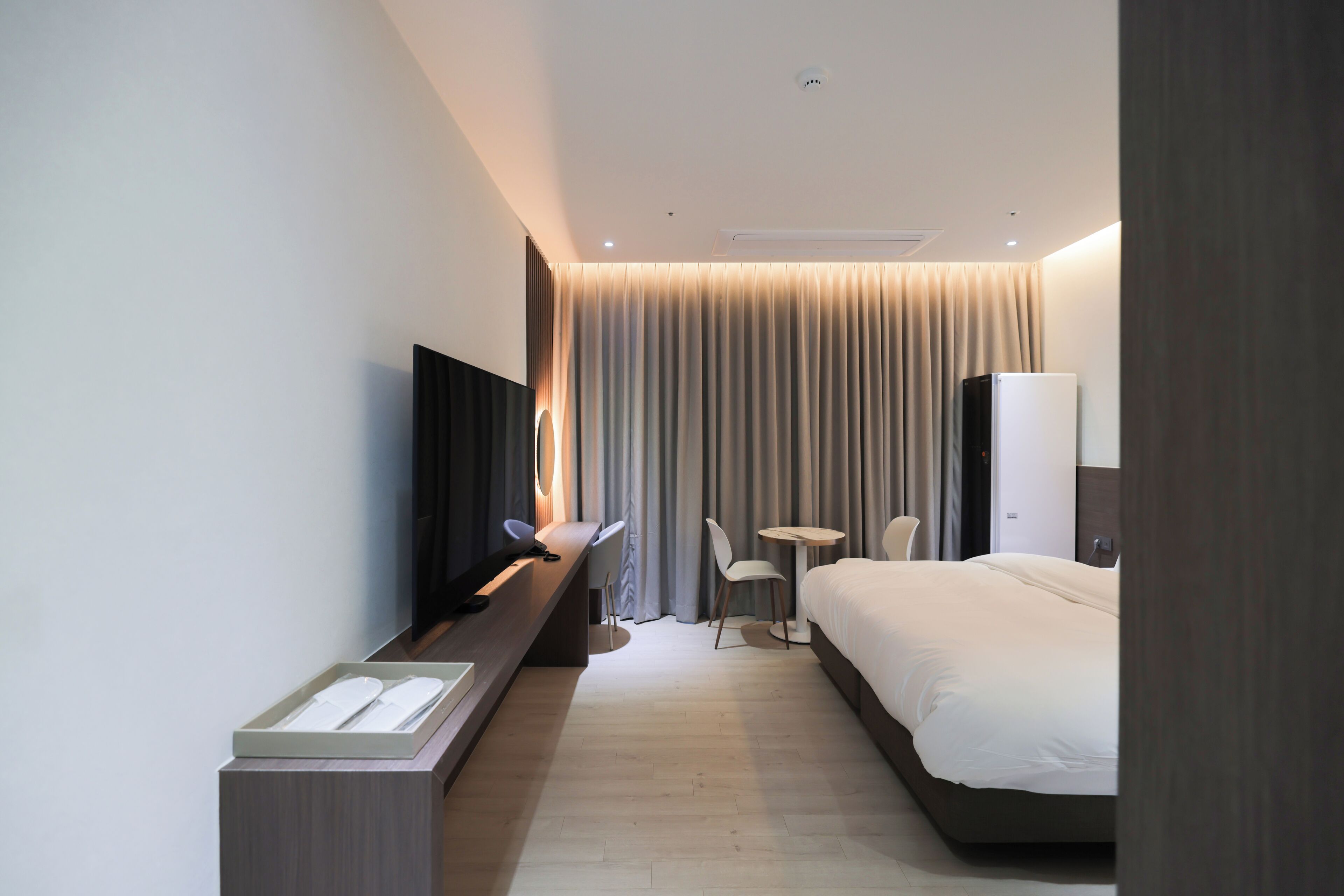 A modern hotel room with a bed, a desk with a flat-screen TV, a small table, and a chair.