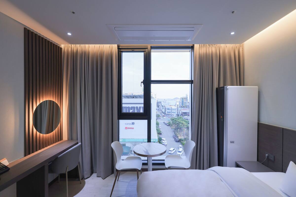 A hotel room with a bed, a desk with a chair, a small table, a TV, and a window with curtains.