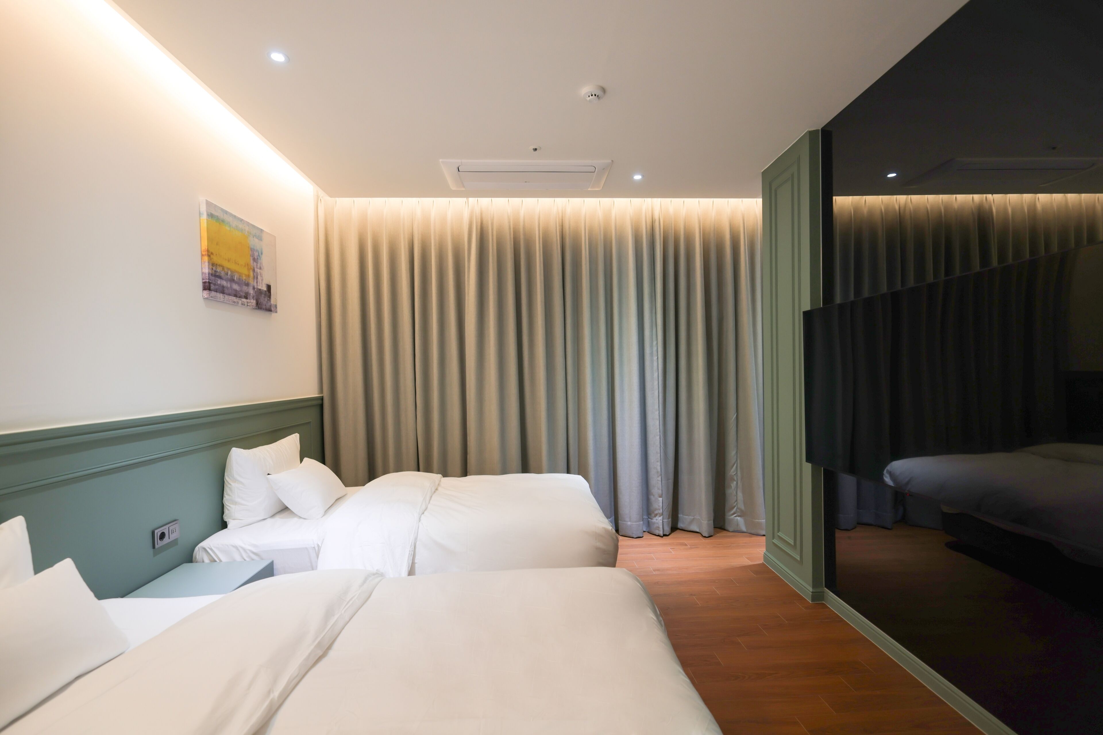 A hotel room with two beds, a large window with curtains, a wall-mounted air conditioner, and a painting.