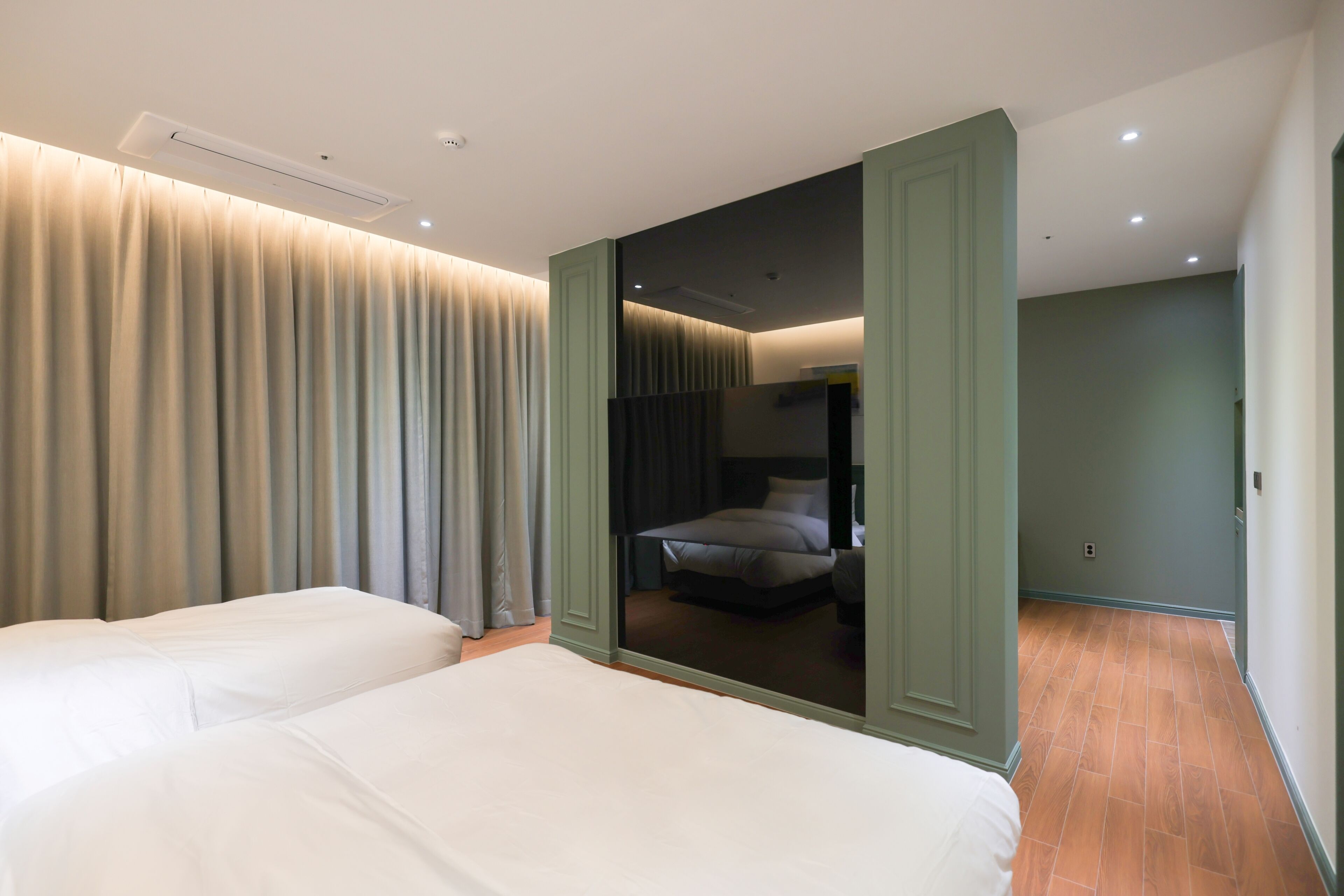 A hotel room with two beds, a large window with curtains, a door leading to another room, and a visible ceiling light.