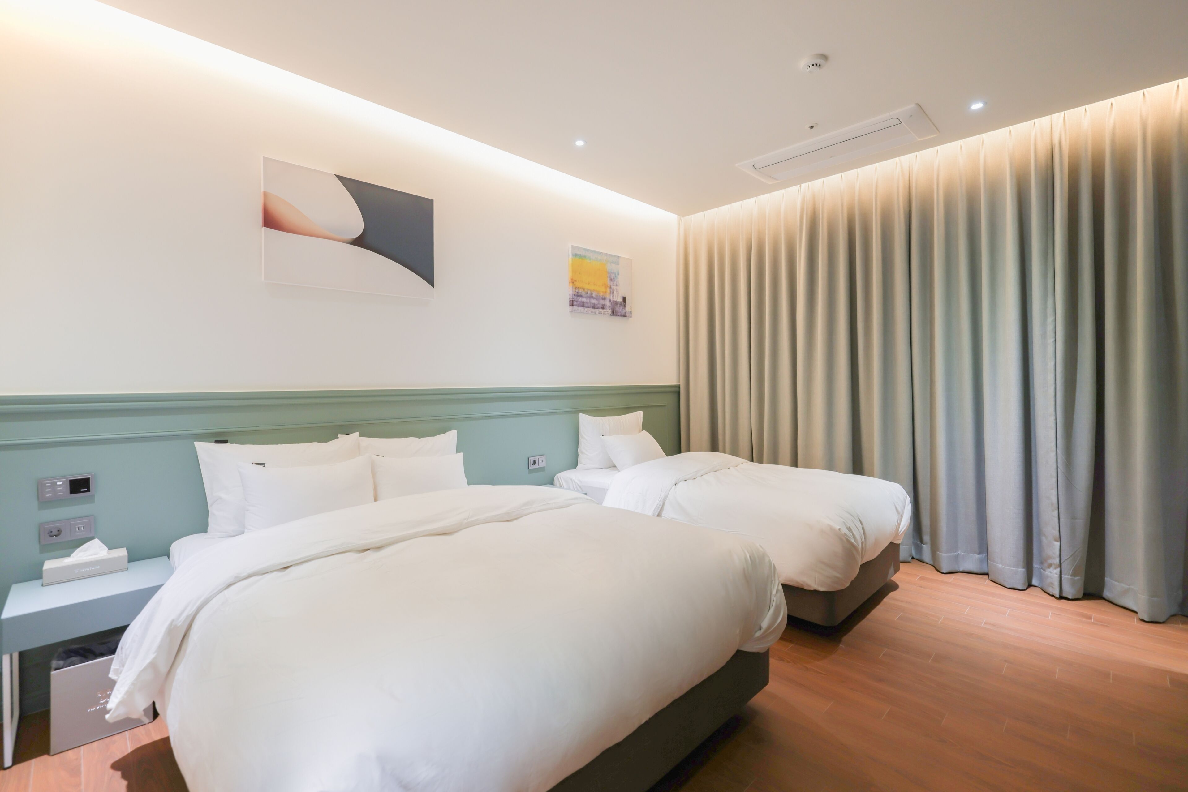 A hotel room with two beds, a nightstand with a lamp, a wall-mounted artwork, and a large window with curtains.