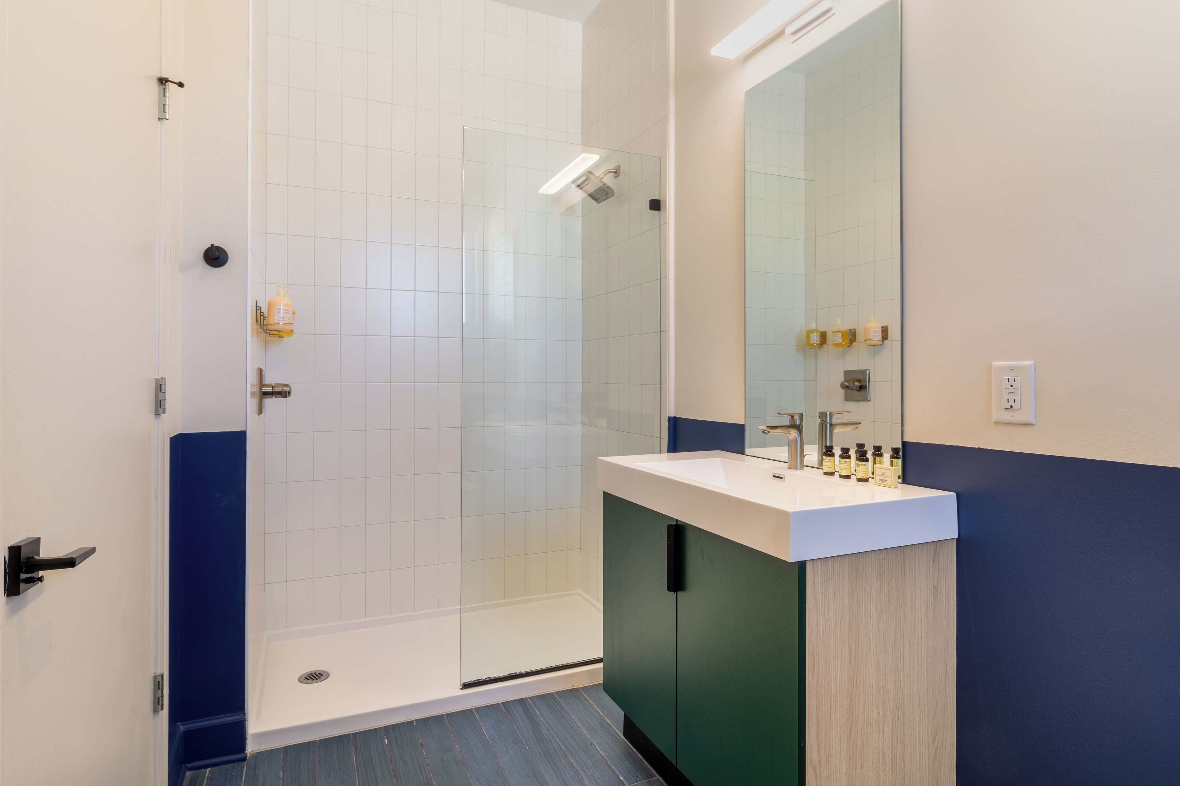 Un baño moderno con cabina de ducha de vidrio, un lavabo blanco y un mueble verde, y un espejo grande.