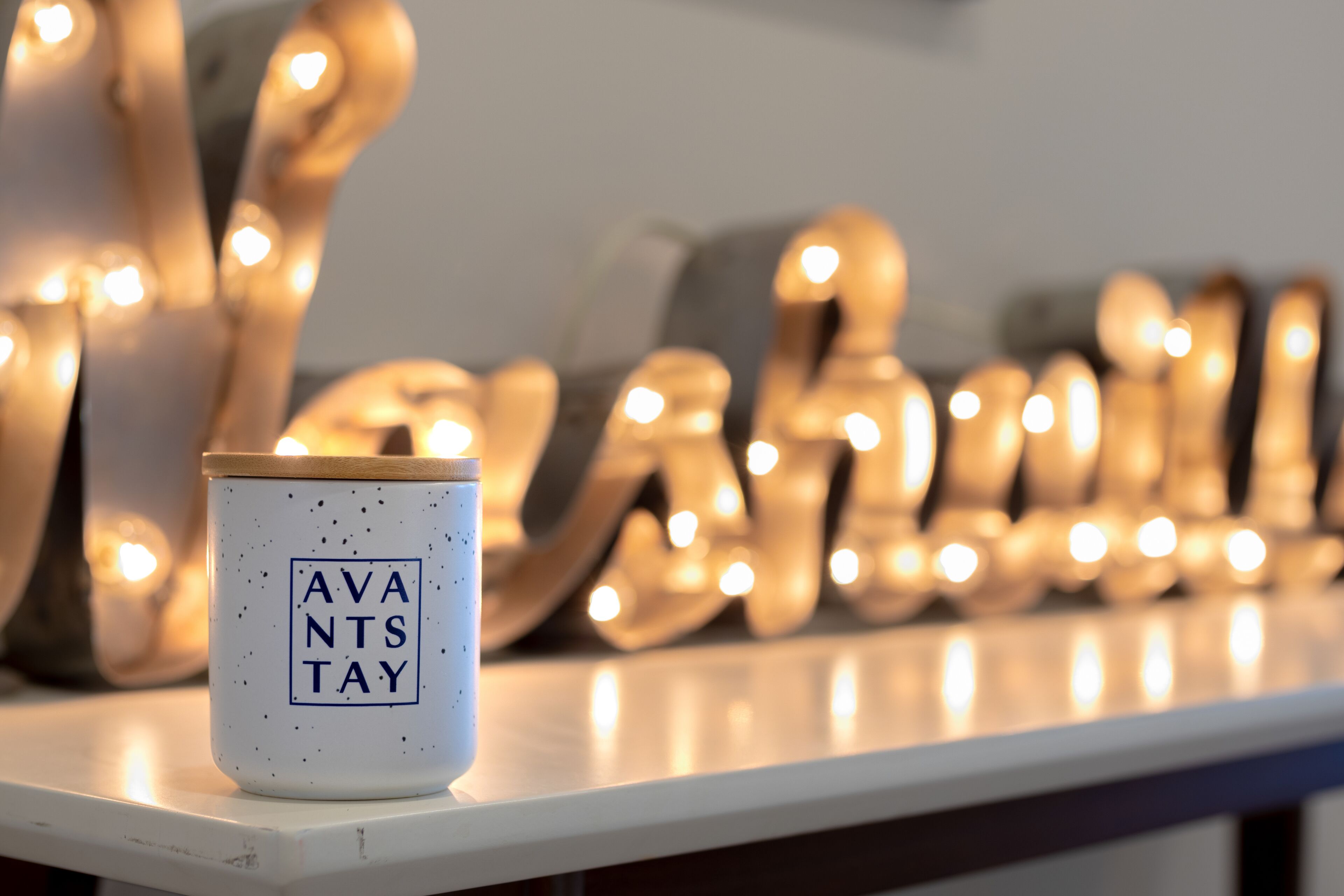 Una taza de cerámica blanca con el texto "AVANTS TAY" sobre una estantería blanca, rodeada de cálida luz de velas.