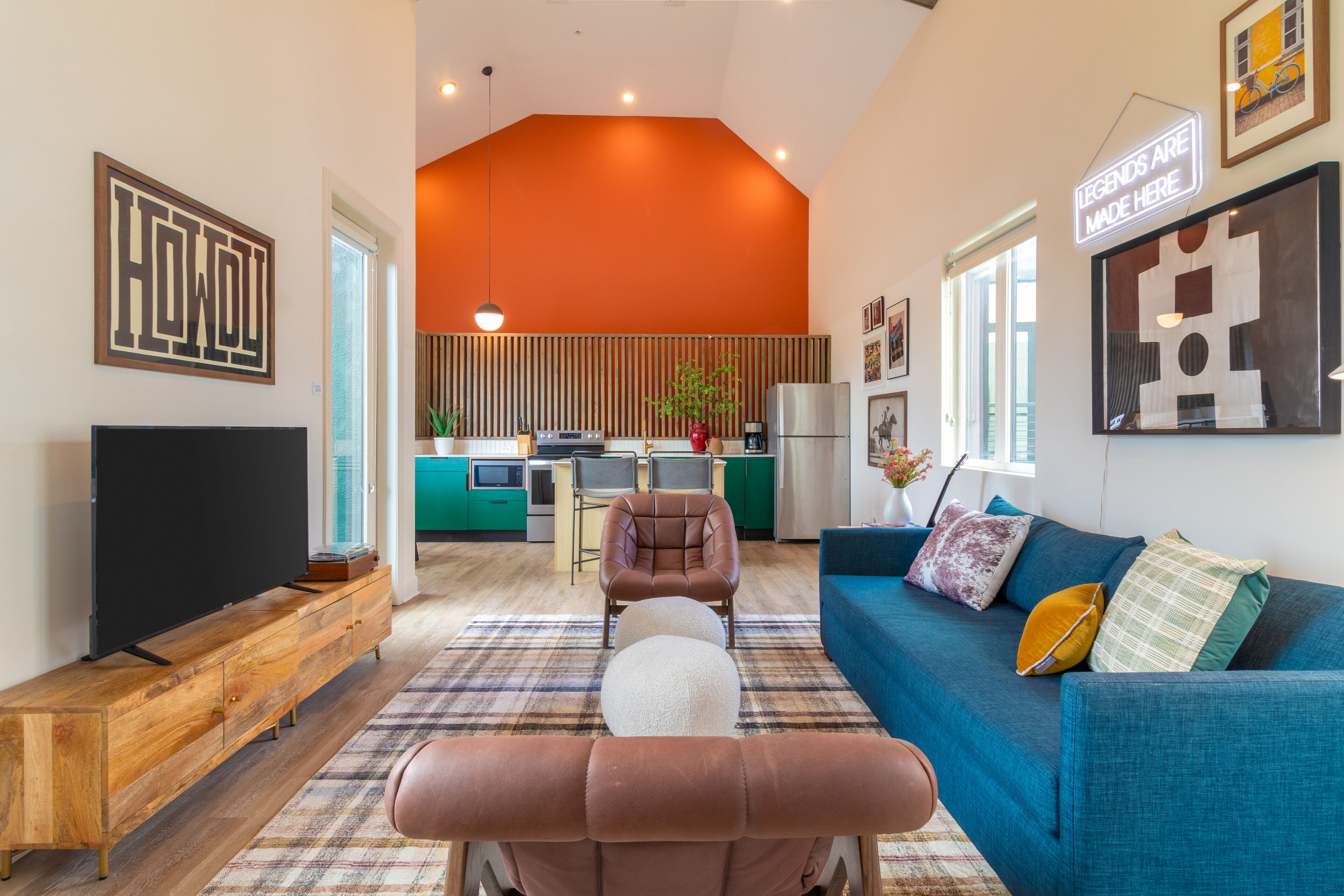 A modern living room with a television, sofa, and ottoman, leading to a kitchen with green cabinets and a dining area.