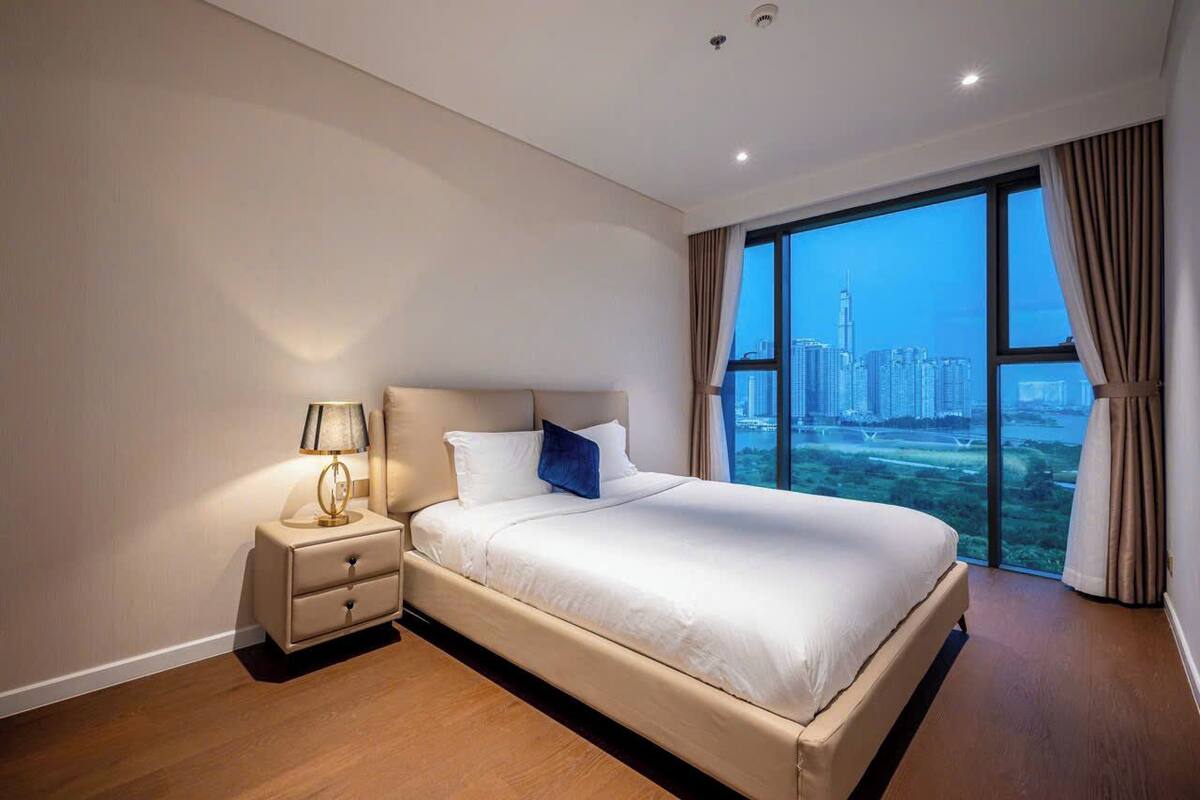 Habitación de hotel moderna con una cama grande, una mesita de noche y vistas a un paisaje urbano a través de un ventanal.