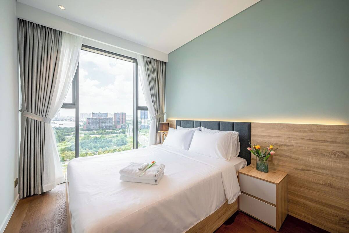 Habitación de hotel con una cama grande, una mesita de noche con un florero y vistas de la ciudad a través de amplios ventanales.