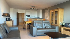 A modern living room with a grey leather sofa, a coffee table, and a dining area with a yellow tablecloth. There is a kitchen in the background with white cabinets and a wooden cabinet with glass doors. - Apartment in Nieuwpoort with Sea Views (Nieuwpoort)