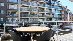 A modern apartment building with multiple balconies, a dining area with a round table and chairs, and a view of a street with cars and a shop named "Boekhandel".