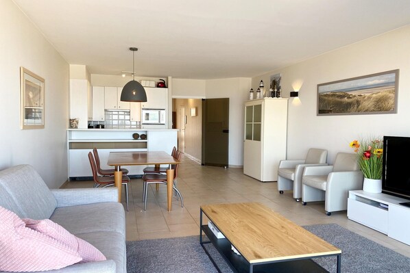 A modern living room with a sofa, coffee table, and TV. There is a dining area with chairs and a table. A kitchen is visible in the background. - Apartment in Nieuwpoort near the Beach (Nieuwpoort)